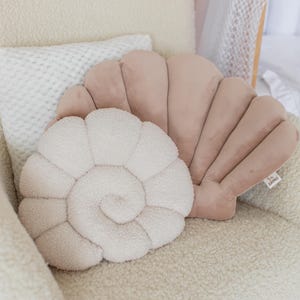 Könnte beinhalten: Zwei dekorative Muschelkissen in Hellrosa und Creme. Ein Kissen ist spiralförmig, das andere muschelförmig. Sie liegen auf einem cremefarbenen, strukturierten Stuhl. Die Kissen sind aus weichem Material.