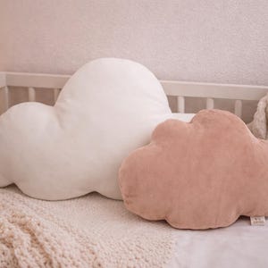 May include: Two cloud-shaped cushions in a nursery setting. One cushion is white, the other is a dusty rose colour. A white cot, a knitted blanket, and a stuffed bunny are also visible. The cushions are soft and plush.