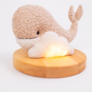 Puede incluir: Una luz de noche de ballena de peluche beige con vientre blanco, sobre una base de madera redonda. Una luz en forma de nube brilla delante de la ballena. La luz de noche está diseñada para una guardería.