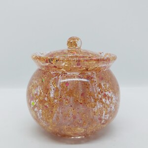 May include: A small, round, lidded jar with a clear, glossy finish. The jar is filled with gold leaf and flecks of pink and green glitter. The lid has a small, round knob. The jar is likely used for storing small items.