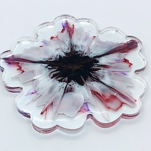 May include: A flower-shaped coaster with a clear, scalloped edge. The coaster features a white base with a dark brown center and white, red, and purple petal-like designs. The coaster is made of resin.