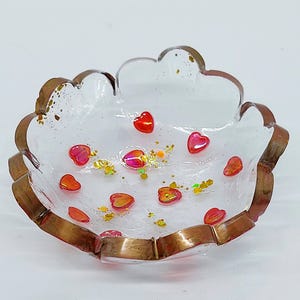 May include: A clear, heart-shaped trinket dish with a bronze-colored rim. The dish is filled with small red and orange heart-shaped decorations and gold glitter. The dish is a decorative item.