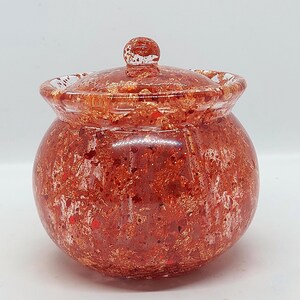 May include: A small, round, lidded jar with a reddish-brown, speckled design. The jar has a clear, rounded lid with a small knob on top. The jar's body is a similar color, with a textured, speckled appearance. The jar is likely made of glass or a similar material.