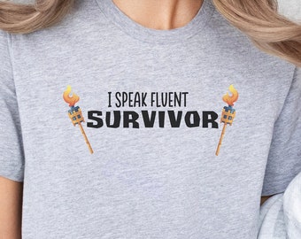 May include: A gray t-shirt with the text "I speak fluent Survivor" and two images of torches on either side of the text.