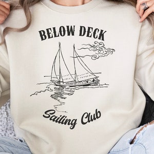 May include: A cream-colored sweatshirt with the text "Below Deck Sailing Club" and a black and white illustration of a sailboat.
