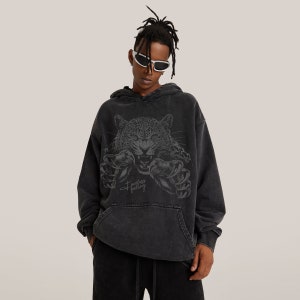 Può includere: Felpa con cappuccio nera con un aspetto vissuto. La parte anteriore presenta una grafica di un giaguaro ringhiante con gli artigli allungati. Il testo "Fearless Feeling" è stampato sotto la grafica.