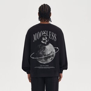 May include: Black sweatshirt with a graphic print of a cherub sitting on a moon with a ring around it. The text "MOONLESS" is printed above the moon and the date "21/07/1969" is printed below the moon.