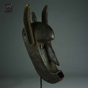 Puede incluir: Una máscara de madera tallada de color marrón oscuro con una forma animal estilizada. La máscara presenta orejas prominentes, un hocico largo y una abertura bucal rectangular. La máscara está montada sobre un soporte contra un fondo neutro, mostrando sus intrincados detalles y artesanía.