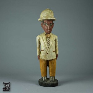 Op de afbeelding: Een houten beeldje van een persoon die een wit pak, een bruine broek en een witte pith-helm draagt. Het beeldje heeft een geschilderd gezicht met zwarte ogen en een rode mond.