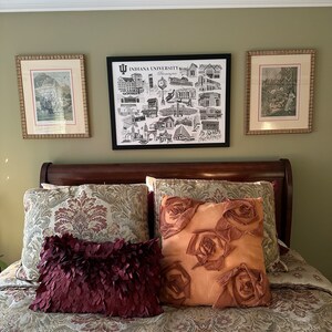 May include: A bedroom with a green wall and a wooden headboard. There are three framed pictures on the wall, one of which is a black and white illustration of Indiana University buildings. The bed is made with a patterned comforter and pillows, including a burgundy pillow with a ruffled texture and a tan pillow with a rose design.