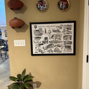May include: A framed black and white print of Indiana University Bloomington campus buildings and landmarks. The print is surrounded by a black frame and has a white mat. The print is hanging on a beige wall with two footballs and two framed pictures of athletes.