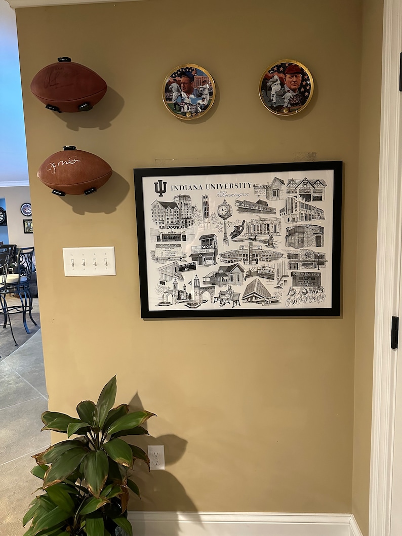 May include: A framed black and white print of Indiana University Bloomington campus buildings and landmarks. The print is surrounded by a black frame and has a white mat. The print is hanging on a beige wall with two footballs and two framed pictures of athletes.