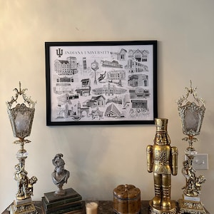 May include: A black and white framed print of Indiana University Bloomington landmarks, a gold nutcracker figurine, and two gold candle holders with crystal accents on a marble surface.