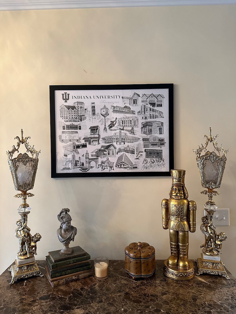 May include: A black and white framed print of Indiana University Bloomington landmarks, a gold nutcracker figurine, and two gold candle holders with crystal accents on a marble surface.
