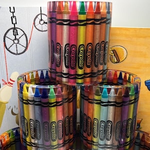 Op de afbeelding: Drie doorzichtige, cilindervormige containers gevuld met kleurrijke Crayola-kleurpotloden. De kleurpotloden zijn verticaal gerangschikt en tonen een spectrum aan kleuren. De containers zijn gestapeld, met kunstwerken op de achtergrond.