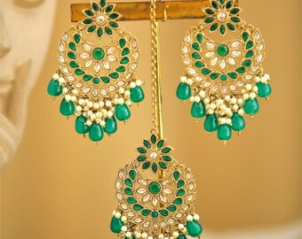 Puede incluir: Juego de joyas doradas con un par de pendientes y un adorno para la cabeza. Cada pieza tiene un diseño floral con detalles verdes y blancos, y cuentas verdes colgantes en forma de lágrima. El conjunto se exhibe en un soporte dorado.