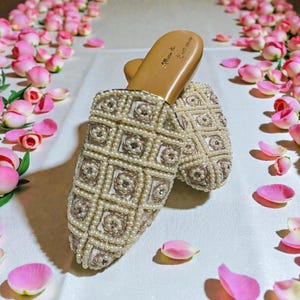 May include: Ivory-colored beaded sandals with a geometric pattern. The sandals are embellished with small, round beads and feature a light brown sole. The sandals are surrounded by pink rose petals, creating a soft, romantic aesthetic.