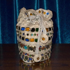 Könnte beinhalten: Ein weißer, mit Perlen besetzter Beutel mit bunten Edelsteinen und silbernen Akzenten. Die Tasche hat einen silbernen Kordelzugverschluss und ist mit Edelsteinen in verschiedenen Formen und Farben verziert, darunter Blau, Gelb und Grün.