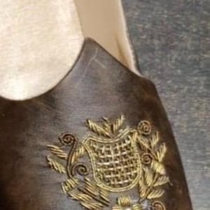 May include: Brown leather shoe with a decorative gold embroidered crest on the vamp. The shoe has a cream-colored interior and a rounded toe. The shoe is likely a loafer or slip-on style.