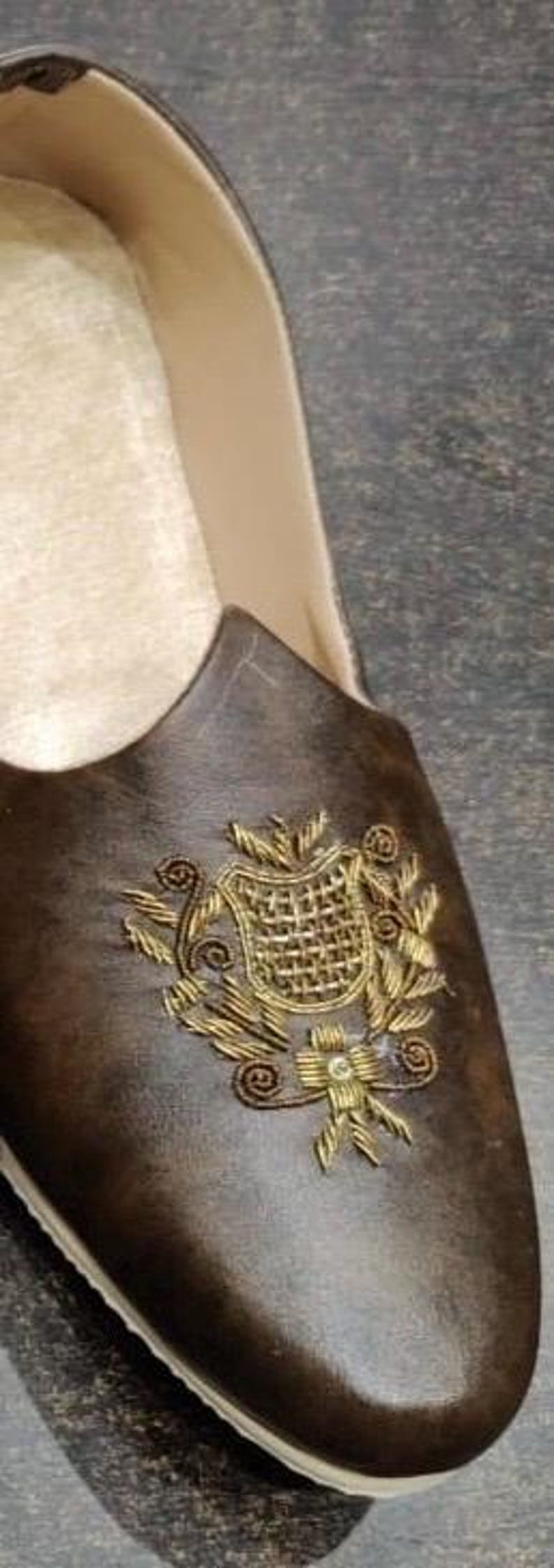 May include: Brown leather shoe with a decorative gold embroidered crest on the vamp. The shoe has a cream-colored interior and a rounded toe. The shoe is likely a loafer or slip-on style.