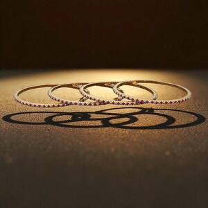 May include: Four gold bangles with red gemstones are displayed against a warm, brown background. The bangles are thin and circular, with the gemstones set along the outer edge. The lighting creates shadows, enhancing the jewelry's elegant design.