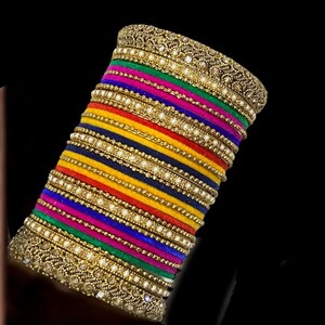 May include: A stack of colorful bangles with gold-toned embellishments. The bangles feature a variety of colors, including green, pink, blue, yellow, and red. The gold accents are detailed with small, clear stones and a textured design.