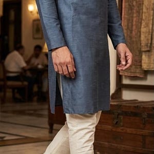 May include: A man in a long-sleeved, slate blue tunic and off-white pants. He wears brown leather loafers with gold embroidery. The outfit is a traditional style, suitable for formal or semi-formal occasions.