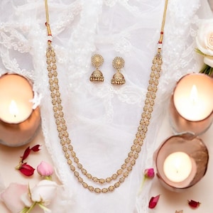 May include: A gold-toned jewelry set featuring a two-strand necklace and matching earrings. The necklace and earrings have a detailed, beaded design. The set is displayed on a white, textured surface, with lit candles and rose petals in the background.