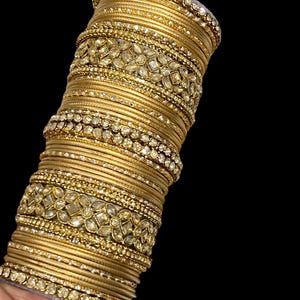 May include: A stack of gold bangles with intricate detailing. The bangles are adorned with sparkling embellishments and a floral pattern. The jewelry is set against a black background, highlighting the gold color and the sparkle of the gemstones.