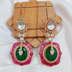 May include: A pair of gold-toned drop earrings. Each earring features a clear, teardrop-shaped stone at the top, followed by smaller clear stones. The lower portion has a green center, surrounded by a pink frame with clear stones.