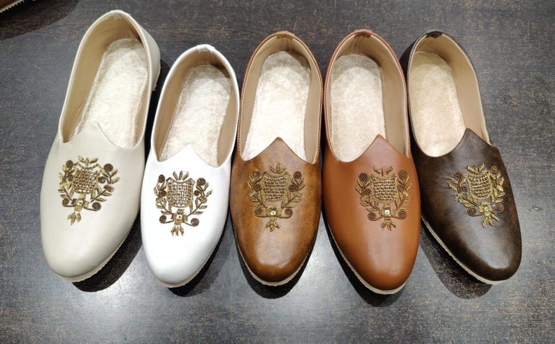 May include: Five pairs of slip-on shoes in cream, white, brown, tan, and dark brown. Each shoe features an embroidered design on the vamp. The shoes have a rounded toe and a low heel, suitable for casual wear.