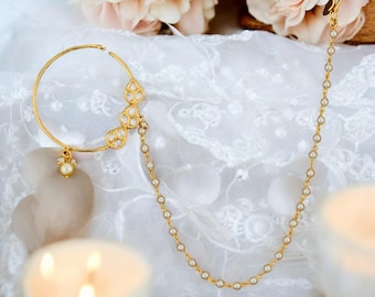 Op de afbeelding: Een goudkleurige neusring met een cirkelvormig ontwerp en een bungelende parel. De ring heeft drie sierlijke, druppelvormige versieringen met heldere stenen. Een delicate ketting van kleine parels loopt van de ring.