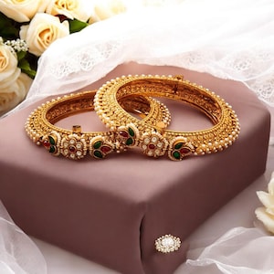 May include: A pair of gold bangles with intricate designs, adorned with white pearls, red and green gemstones. The bangles are on a soft, purple surface.