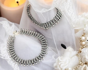 Pode incluir: Um conjunto de colar e pulseira prateados com um design geométrico repetitivo e pequenas pedras transparentes. As joias são exibidas em uma superfície branca texturizada com uma vela acesa e flores brancas ao fundo.