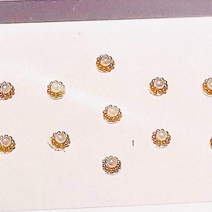 May include: A sheet of small, round, gold-colored bindi stickers with a central pearl-like accent. The stickers are arranged in rows and columns on a white background.