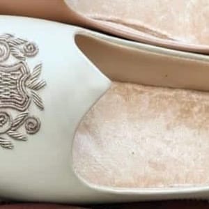 May include: Cream-colored slip-on shoes with a decorative floral design. The shoes have a soft, light brown interior and are made of a smooth material. The shoes are displayed on a wooden surface.