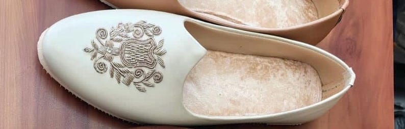 May include: Cream-colored slip-on shoes with a decorative floral design. The shoes have a soft, light brown interior and are made of a smooth material. The shoes are displayed on a wooden surface.