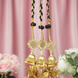 May include: Three ornate gold-colored hair accessories with black braided cords. Each piece features a bell-shaped design with intricate detailing, including small pearls and green accents. The background is a soft pink fabric.