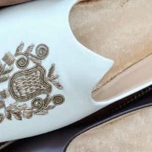 May include: Close-up of three pairs of leather slip-on shoes. One pair is white with gold embroidery, another is dark brown, and the third is a lighter brown. The shoes are arranged on a wooden surface.
