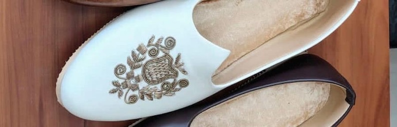 May include: Close-up of three pairs of leather slip-on shoes. One pair is white with gold embroidery, another is dark brown, and the third is a lighter brown. The shoes are arranged on a wooden surface.