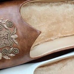 May include: A pair of leather slippers. One slipper is brown with gold embroidery and a soft, beige interior. The other slipper is white with a beige interior. The slippers are designed for indoor use.