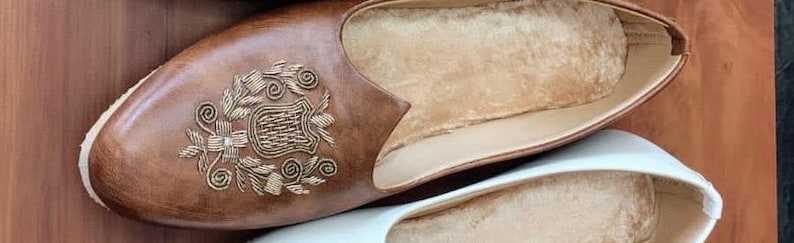 May include: A pair of leather slippers. One slipper is brown with gold embroidery and a soft, beige interior. The other slipper is white with a beige interior. The slippers are designed for indoor use.