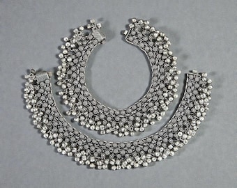 Pode incluir: Duas pulseiras de tornozelo prateadas com detalhes intrincados e pequenas sinetas penduradas. As pulseiras são exibidas sobre um fundo cinzento, mostrando o seu design ornamentado e o seu artesanato. A joalharia foi concebida para ser usada à volta do tornozelo.