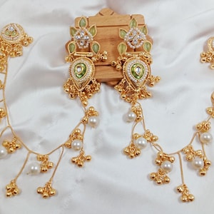 May include: Gold-toned earrings with intricate detailing and dangling pearl accents. The earrings feature a central design with green and white embellishments, and a delicate chain of pearls and gold beads.