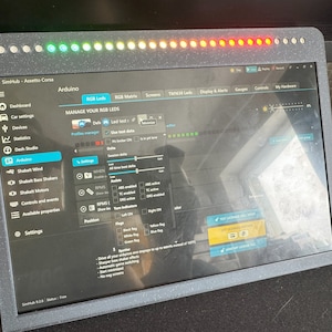 May include: A computer screen displaying a dashboard with the title "SimHub - Assetto Corsa". The dashboard has a menu with options like "Dashboard", "Car settings", "Devices", "Dash Studio", "Arduino", and "Settings". The screen also shows a section for managing RGB LEDs with options for "Led test", "Blue test data", "Session data", and "Position".