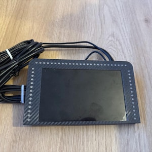 May include: A black electronic device with a screen and a carbon fibre-style frame. The frame is dotted with small, evenly spaced lights. Black cables are attached to the device and bundled with white ties.