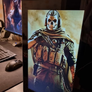 Peut inclure: Un écran d'affichage numérique montrant un personnage de jeu vidéo portant un masque de crâne blanc et un gilet tactique marron. Le personnage tient une arme à feu et porte un motif de camouflage sur ses vêtements.