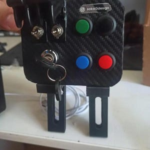 Puede incluir: Un panel negro de fibra de carbono con cuatro botones, dos interruptores de palanca y un interruptor de llave. El panel está montado en dos soportes negros. El texto "sakai3design" está impreso en el panel.