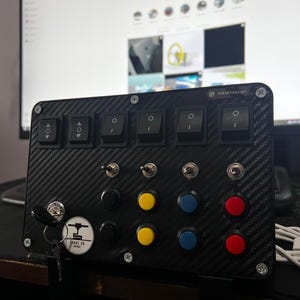 Puede incluir: Un panel negro con un patrón de fibra de carbono, que presenta cuatro interruptores de palanca, seis botones redondos y una llave con un logotipo que dice "Sakal 3D Design".