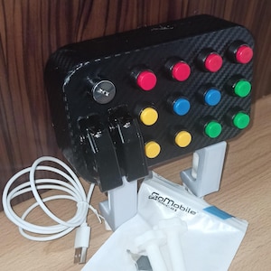 May include: A black gaming controller with a carbon fiber design. It has multiple buttons in red, blue, green, and yellow. The controller is connected to a computer via a USB cable. The controller is labeled "GoMobile GMMC-01".